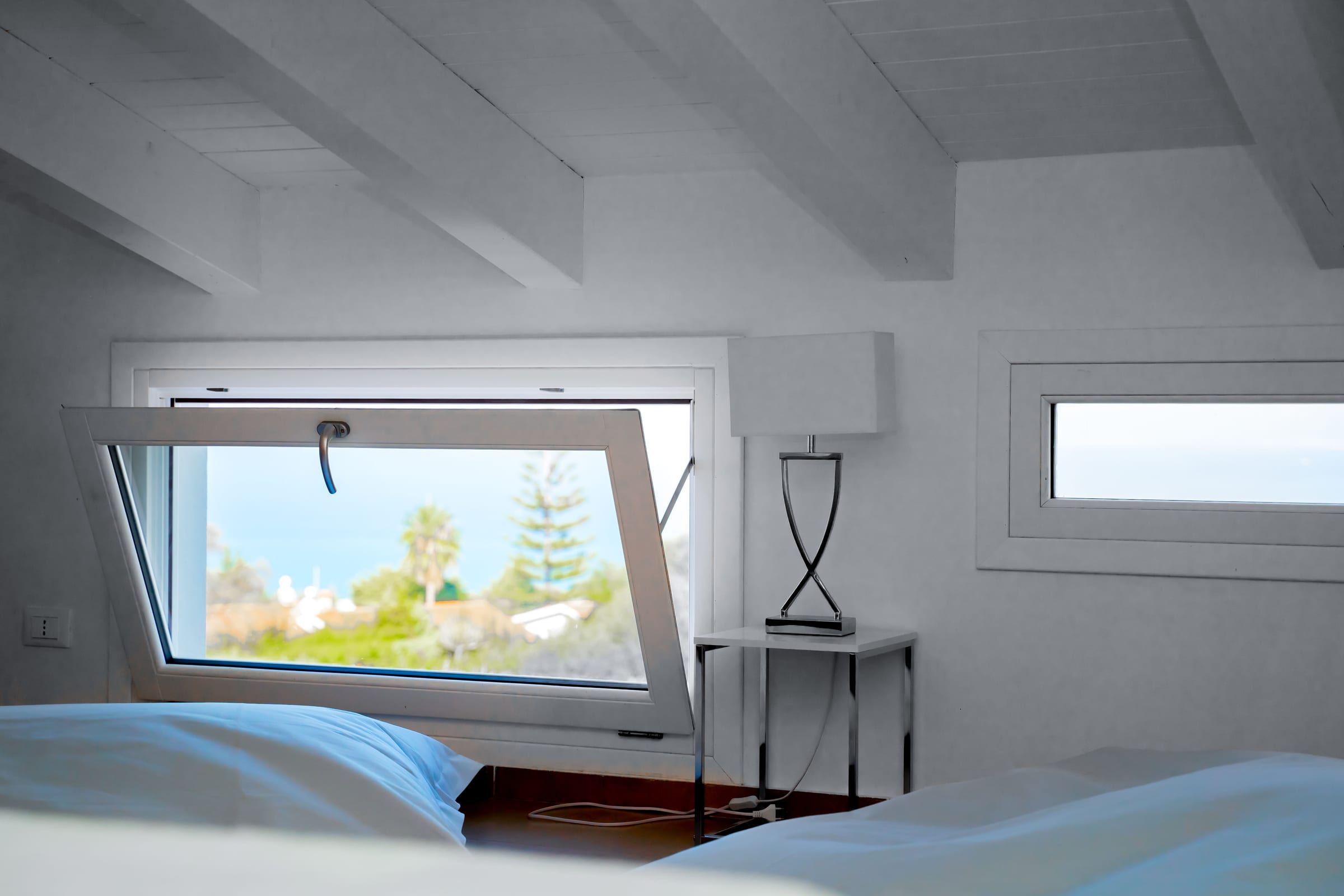 Bedroom under a white beamed ceiling; the tilted window looks out over trees to the sea