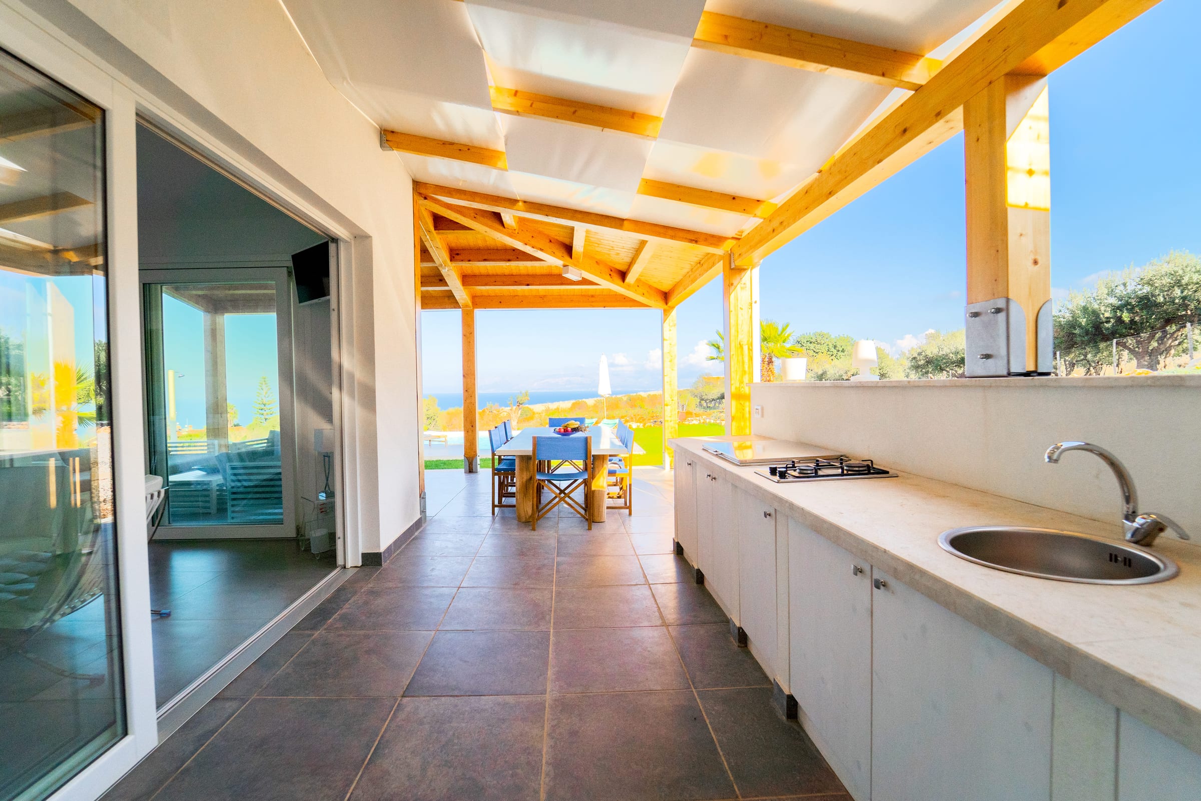 Outdoor kitchen with hob and sink under the wooden pergola, looking past the dining table to the sea