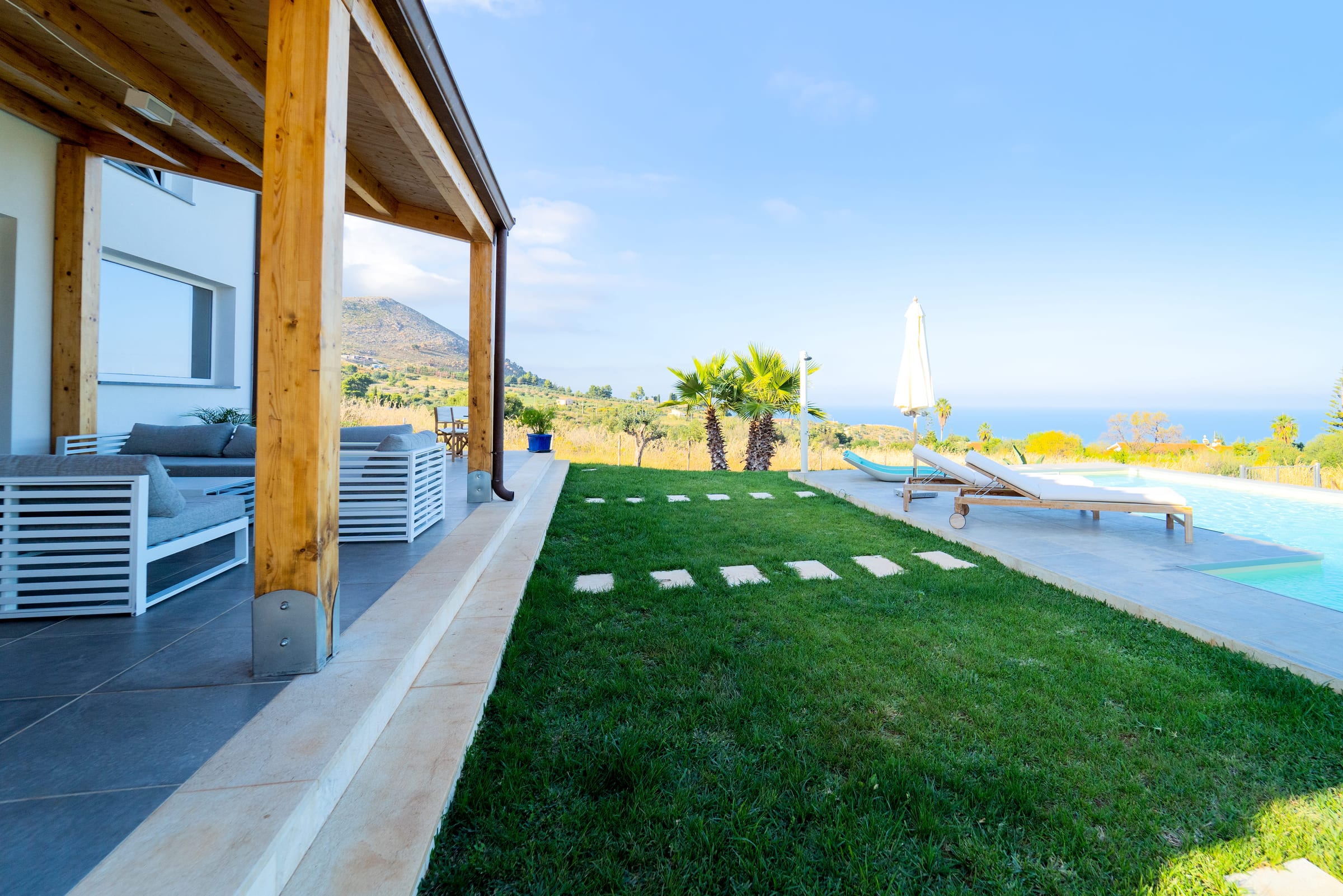 From the covered terrace, stepping stones lead across the lawn to the pool and its sun loungers, with the sea in the distance