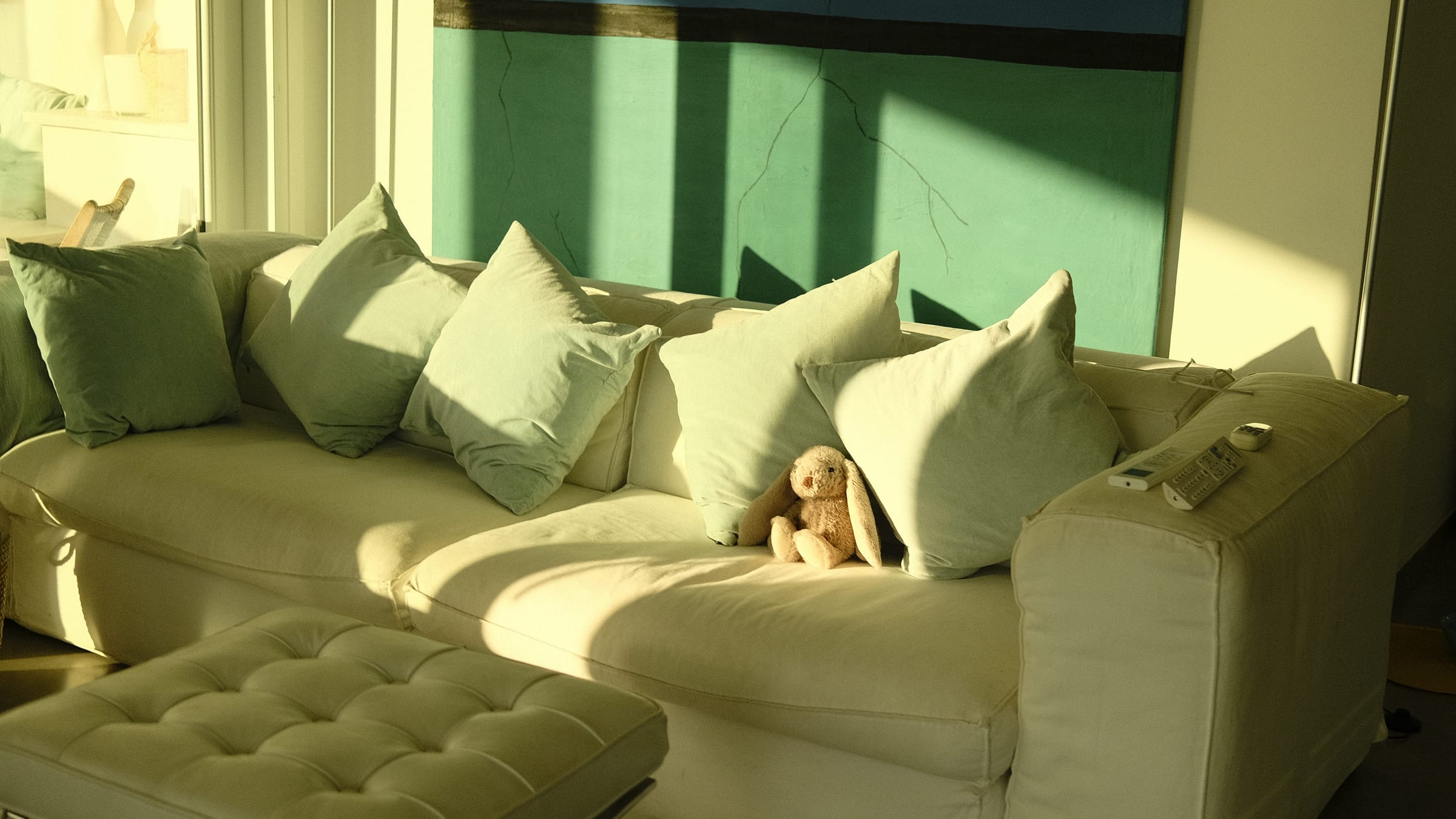 White sofa with pale green cushions and a toy rabbit, a tufted footstool in front and a large wall painting behind, in low sun